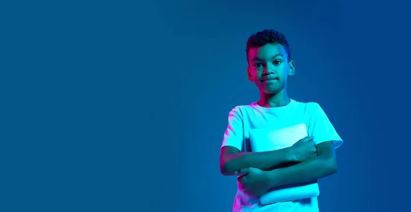 Remote education. Serious little boy, kid in white t-shirt holding tablet and looking at camera isolated over blue background in neon light. Education, childhood, modern lifestyle