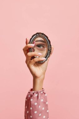 Retro tarzı. Pembe arka planda izole edilmiş kızların gözünün yansıması olan küçük bir ayna tutan kadın el. Klasik moda, güzellik, sanat, yaratıcılık ve reklam kavramı. İnsani duygular, yüz ifadesi