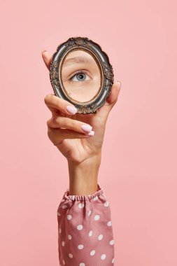 Retro tarzı. Pembe arka planda izole edilmiş kızların gözünün yansıması olan küçük bir ayna tutan kadın el. Klasik moda, güzellik, sanat, yaratıcılık ve reklam kavramı. İnsani duygular, yüz ifadesi