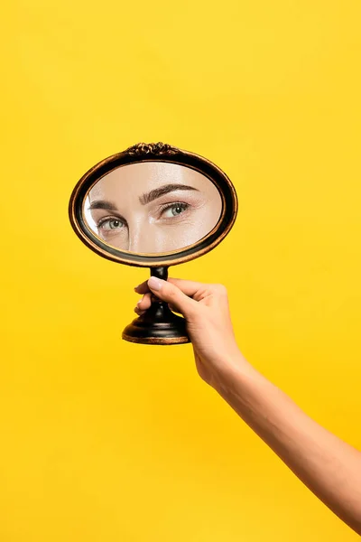 Happy look. Female hand holding mirror with reflection of girls eyes isolated over yellow background. Concept of vintage, retro style, beauty, art, creativity and ad. Human emotions, facial