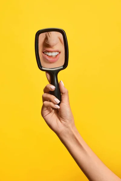 Female hand holding mirror with reflection of womans lips and teeth isolated over yellow background. Concept of vintage, retro style, beauty, art, creativity and ad. Human emotions, facial expression