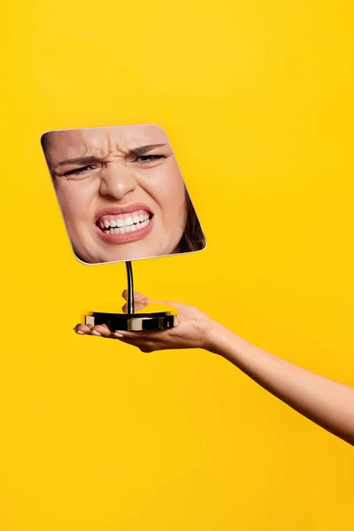 Angry. Female hand holding mirror with reflection of angry girls face isolated over yellow background. Concept of vintage, retro style, beauty, art, creativity, ad. Human emotions, facial expression