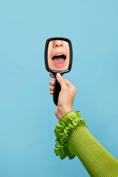 Female hand holding mirror with reflection of shouting girls mouth isolated over blue background. Concept of vintage, retro style, beauty, art, creativity and ad. Human emotions, facial expression