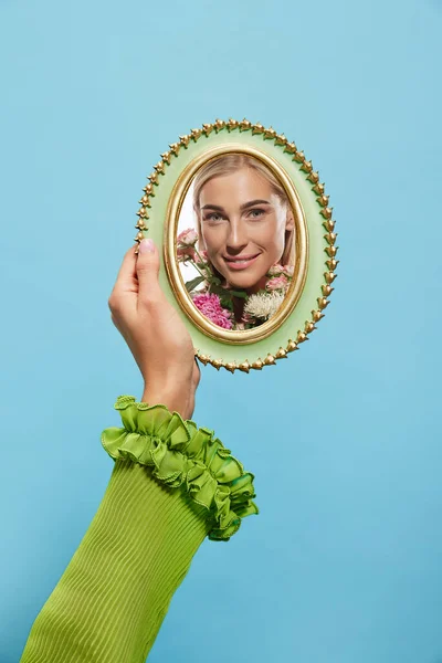 Female hand holding mirror with reflection of girls face isolated over blue background. Concept of vintage, retro style, beauty, art, creativity and ad. Human emotions, facial expression