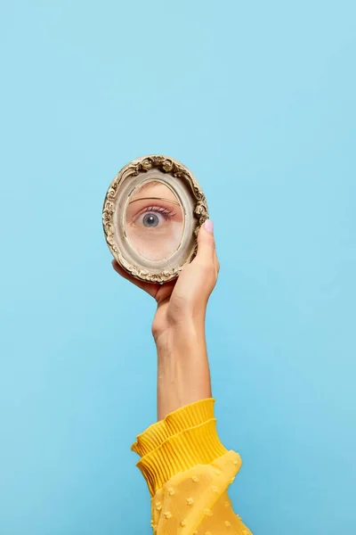 Eyesight. Female hand holding small round mirror with reflection of girls eye isolated over blue background. Concept of vintage fashion, beauty, art, creativity and ad. Human emotions, facial