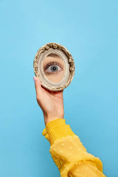 Eyesight. Female hand holding small round mirror with reflection of girls eye isolated over blue background. Concept of vintage fashion, beauty, art, creativity and ad. Human emotions, facial