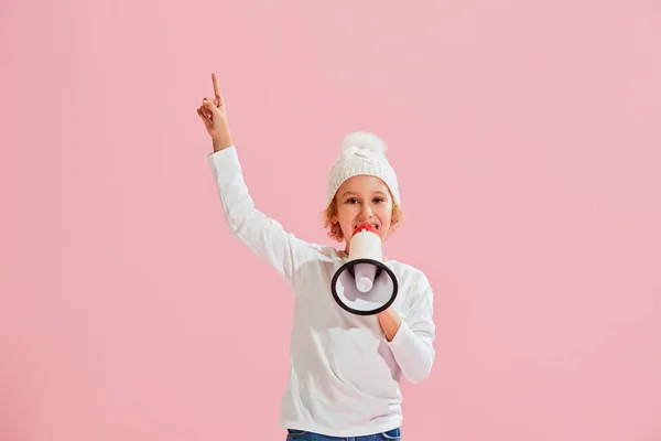 News. Studio shot of little girl, kid in casual style clothes and warm hat shouting at megaphone isolated over pink background. Fashion, winter holidays, vacations. Concept of emotions, dreams, news