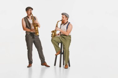 Retro tarzı kıyafetler giymiş iki genç müzisyen beyaz stüdyo arka planında saksafon çalıyor. Müzik kavramı, hobi, festival, sanat, ilham