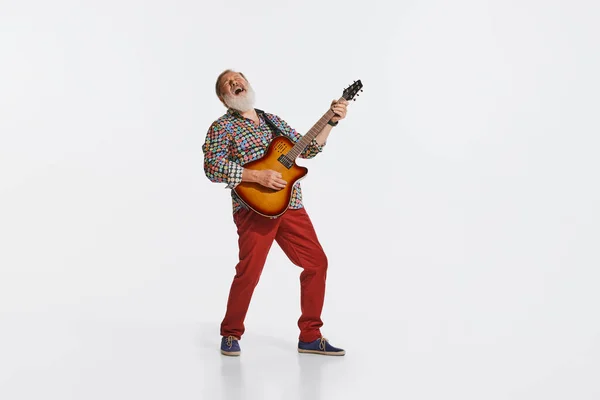 Solo performance. Senior man, musician wearing retro style clothes playing guitar like rockstar isolated on white background. Vintage fashion, music, art, emotions, music festival concept.