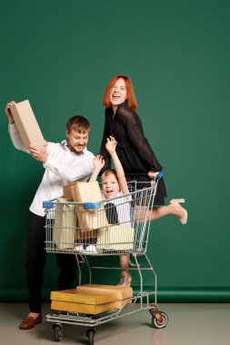 Alışverişe. Neşeli aile, anne, baba ve oğlu alışveriş çantaları ve kutularıyla yeşil arka planda izole edilmiş alışveriş arabalarına biniyorlar. Satış kavramı, Kara Cuma, indirim, duygular..