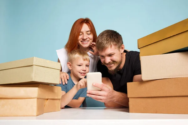 Happy family, parents and kid among cardboard boxes using smartphone and making online goods order. Black Friday, big sales, leisure activity concept. Unbox package, post shipping delivery