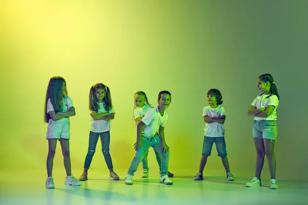 Hip-hop dancing. Group of children, little girls and boys in sportive casual style clothes dancing in choreography class isolated on green background in yellow neon. Concept of music, fashion, art