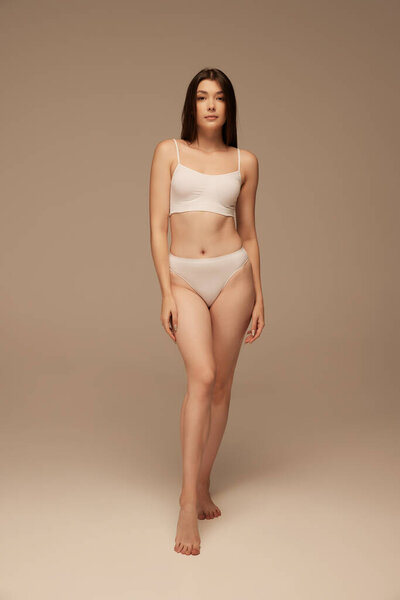 Полнометражный портрет молодой красивой женщины, позирующей в белых трусах, изолированный на темно-бежевом фоне студии. Концепция красоты, ухода за телом и кожей, здоровья, пластической хирургии, косметики, спорта