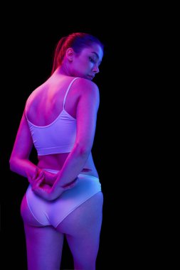 Beyaz iç çamaşırlarıyla poz veren genç, güzel bir kadın. Siyah arka planda, neon ışıkta izole edilmiş. Yan görüş. Sırtın güçlü. Güzellik, vücut ve cilt bakımı, sağlık, estetik cerrahi, kozmetik