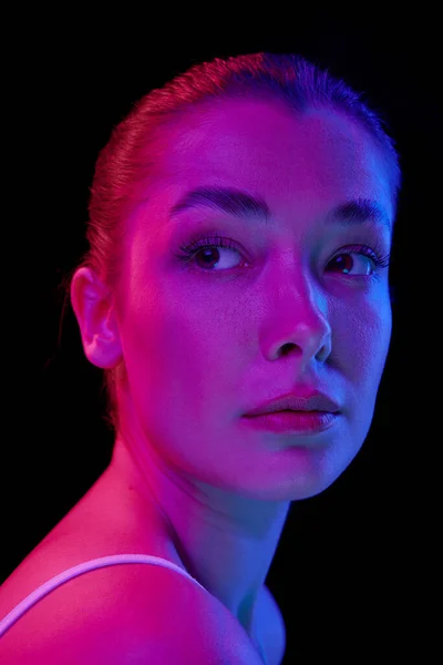 Cose-up portrait of young beautiful woman with clear spotless face looking away, posing isolated over black background in neon light. Concept of beauty, skin care, health, plastic surgery, cosmetics