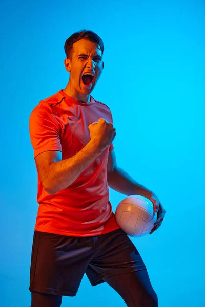 Happy emotions. Young male volleyball player with ball shouting isolated on blue studio background in neon light. Concept of summer sports, gym, team sport, challenges.