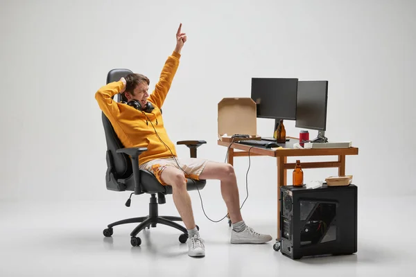 Gamer. Young man in homewear sitting at home, playing computer games isolated over grey background. Business, studying, education, youth, remote workplace concept. Copy space for ad, text