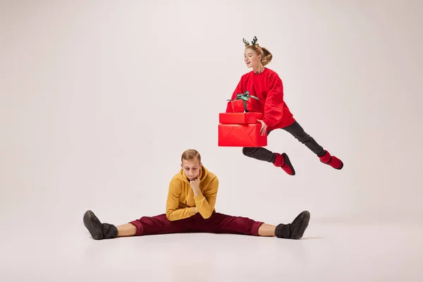 Christmas deer with presents. Flexible dancers, young happy couple in warm winter clothes dancing with festive gift boxes over grey background. Happy New year, holidays, 2023, party concept