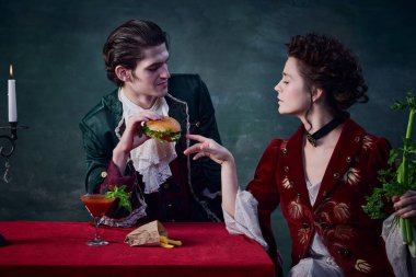 Koyu yeşil arka planda yaşayan vampirlerin portresi. Hamburgerli adam, kerevizli kadın. Sağlıklı ve abur cubur. Cadılar Bayramı kavramı, gizem, gotik sanat, fantezi, gerçeküstü karakterler.