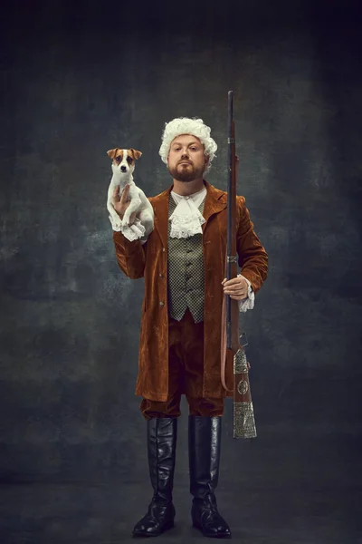 Hunter with doggy. Portrait of young man in image of nobleman in vintage brown hunting suit and white wig with old hunting rifle isolated over dark retro background. Art, fashion, traditions, male