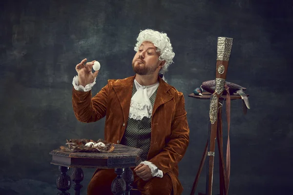 Vintage portrait of young man in brown vintage suit and white wig like medieval royal hunter isolated on dark background. Comparison of eras concept, renaissance style.