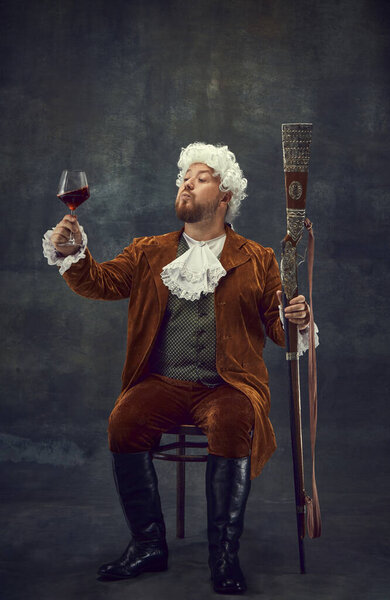 Tasting red wine. Vintage portrait of young man in brown retro suit and white wig like medieval royal hunter isolated on dark background. Comparison of eras concept, renaissance style.