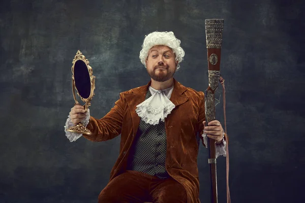 Looking at mirror. Vintage portrait of young man in brown vintage suit and white wig like medieval royal hunter isolated on dark background. Comparison of eras concept, renaissance style.
