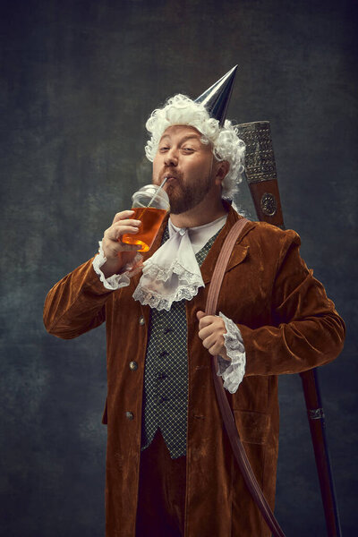 Sweet drinks. Smiling young man in brown vintage suit and white wig like medieval royal hunter drinking lemonade isolated on dark background. Comparison of eras concept, renaissance style.