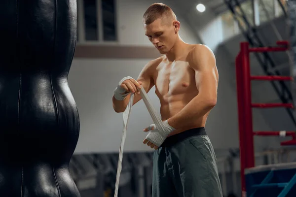 Male boxer wrapping his hands before fighting at sports gym, indoors. Close-up of young athlete getting ready for fight. Sport concept. Athlete looks serious, motivated and concentrated