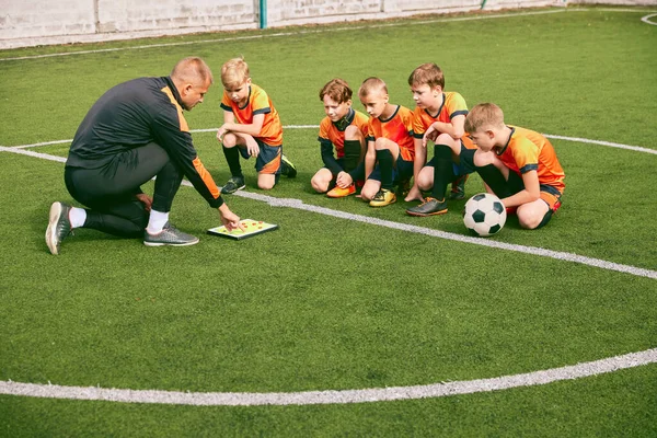 Football training. Soccer coach explaining game rules and strategy using tablet, map. Sports junior team sitting on grass pitch with trainer. Concept of sport, achievements, studying, goals, skills