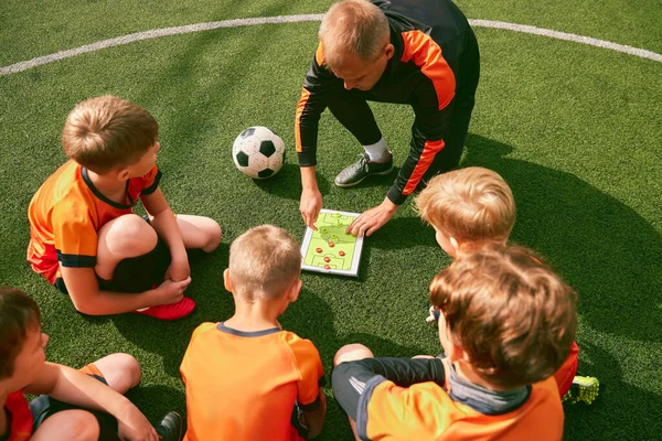 Football training. Soccer coach explaining game rules and strategy using tablet, map. Sports junior team sitting on grass pitch with trainer. Concept of sport, achievements, studying, goals, skills