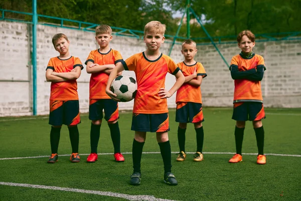 チームメイト 草の野球場で一緒に立っているジュニアサッカーチームのアスレチック少年 オレンジブラックのキットとブーツでサッカー選手 スポーツ 成功とスキルの概念 — ストック写真