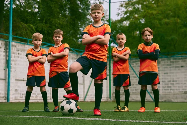 Teammates. Athletic boys in junior soccer team standing together at grass sport field. Football players in orange-black kits and boots. Concept of sport, studying, achievements, success and skills