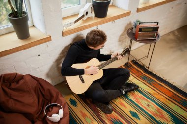Evde yerde oturup gitar çalmaya meyilli genç bir adamın hava manzarası. Hobiler ve boş zaman aktiviteleri. Uzaktan eğitim, online dersler. Sıcak bir atmosfer. Musk, sanat ve benlik kavramı