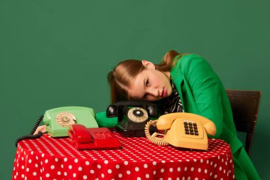 Zorunlu bekleyiş. Retro objeler ve modern gençlik. Eski tarz kıyafetleri olan genç bir kız eski tip telefonları olan bir masada oturuyor. Tamamlayıcı moda renkler. Eski, eski, 80 'ler, 70' ler tarzı.