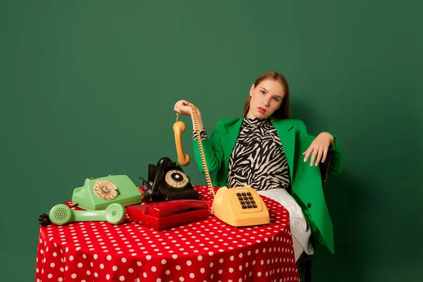 流行艺术摄影 复古物品和现代青年 穿着复古风格衣服的年轻姑娘坐在餐桌旁 手里拿着老式电话 补充流行色彩 80年代 70年代风格 — 图库照片