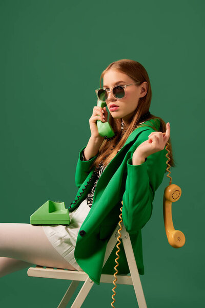 Creative portrait of young without emotions girl in green jacket sitting on chair with retro phone isolated over green background. Vivid style, beauty, queer, freak, fashion concept. Copy space for ad