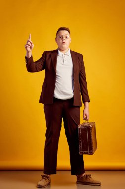 Sarı arka planda izole edilmiş kameraya bakan bavullu, eski moda iş kostümü giymiş genç bir çocuk. Klasik moda, eğitim, üniversite. İnsani duygular, yüz ifadesi, reklam kavramı.