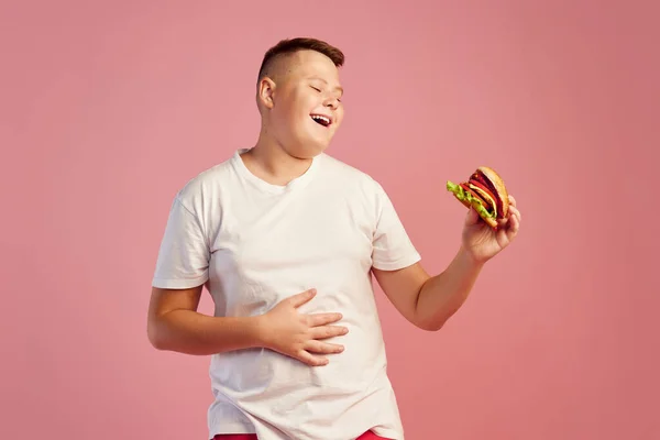 티셔츠를 배경으로 맛있는 햄버거를 맛보는 귀여운 소년의 반감기 패스트푸드 광고용 — 스톡 사진