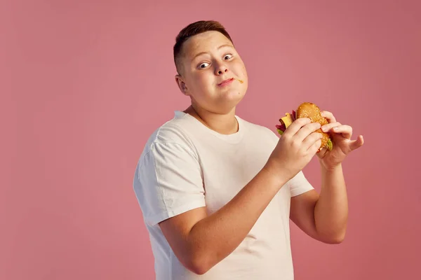 티셔츠를 배경으로 맛있는 햄버거를 맛보는 귀여운 소년의 반감기 패스트푸드 광고용 — 스톡 사진