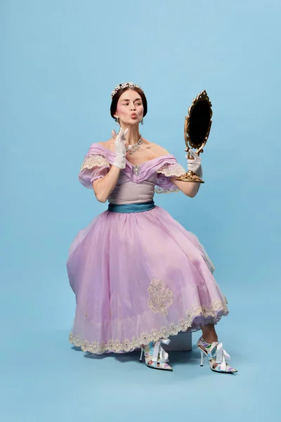 Self loving. Beautiful charming woman in lilac color vintage dress as royal person, princess looking in mirror at herself on blue background. Concept of comparison of eras, modernity and renaissance.