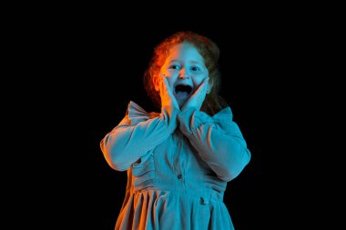 Şaşırmış ve heyecanlı çocuk. Şenlikli elbiseler giyen sevimli kızıl saçlı kız karanlık arka planda, neon ışıkta izole edilmiş. Çocukların duyguları, yüz ifadeleri, moda.