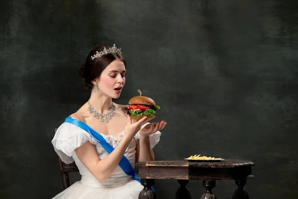 Wow. Young beautiful woman, royal person, queen or princess in white medieval outfit tasting burger on dark background. Concept of comparison of eras, modern, fashion, beauty.