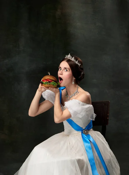 Very tasty. Emotional woman, royal person, queen or princess in white medieval outfit eating burger on dark background. Concept of fast food, facial expressions, eras comparison, fashion, beauty.