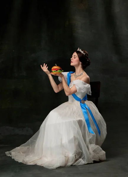 Refined taste. Young beautiful queen or princess in white medieval outfit eating burger on dark background. Concept of comparison of eras, fast food, fashion, beauty and ad