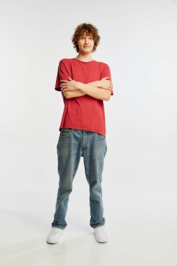 Kollarını çapraz yapmış utangaç genç bir adam beyaz arka planda izole edilmiş. İnsan duyguları, gençlik, satış, reklam. Model, aktör kırmızı pamuklu tişört ve kot pantolon giyiyor