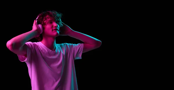 Soul music. Happy young man, student in white tee and headphones isolated on dark background in neon light. Concept of emotions, facial expression, youth, aspiration, sales and ad