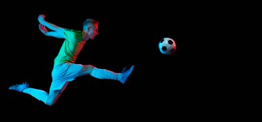 Sıçrayan, futbol topunu tekmeleyen. Genç aktif futbolcunun hareketli görüntüsü karanlık arka planda neon ışıkta izole edilmiş. Spor kavramı, hedefler, rekabet, hobi, reklam.