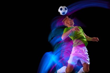 Sıçrayan, futbol topunu tekmeleyen. Genç aktif futbolcunun hareketli görüntüsü karanlık arka planda neon ışıkta izole edilmiş. Spor kavramı, hedefler, rekabet, hobi, reklam.