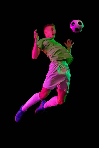 Jumping, kick football ball. Dynamic shot of young active football player in action isolated on dark background in neon light. Concept of sport, goals, competition, hobby, ad.